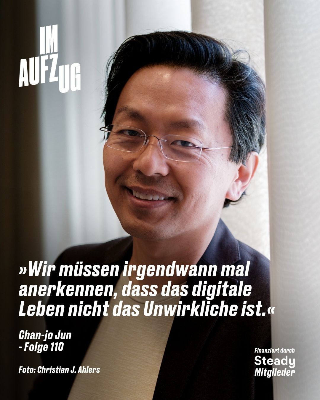 Das Bild zeigt ein Porträt von Chan-jo Jun für den Podcast „IM AUFZUG“ (Folge 110). Der Mann ist im Nahporträt zu sehen, hat schwarzes, leicht leicht nach hinten gestyltes Haar, trägt eine randlose Brille und lächelt freundlich direkt in die Kamera. Er trägt ein helles T-Shirt unter einem dunklen Sakko und lehnt sich leicht an ein Fenster mit hellem Vorhang. Auf der linken Bildseite befindet sich ein großes, weißes Zitat in kursiver Schrift: „Wir müssen irgendwann mal anerkennen, dass das digitale Leben nicht das Unwirkliche ist.“ Unter dem Zitat stehen sein Name und die Episodennummer „Chan-jo Jun - Folge 110“. Das Podcast-Logo „IM AUFZUG“ befindet sich oben links. Am unteren Bildrand stehen zwei kleine Hinweise: links der Bildnachweis „Foto: Christian J. Ahlers“ und rechts der Text „Finanziert durch Steady Mitglieder“.